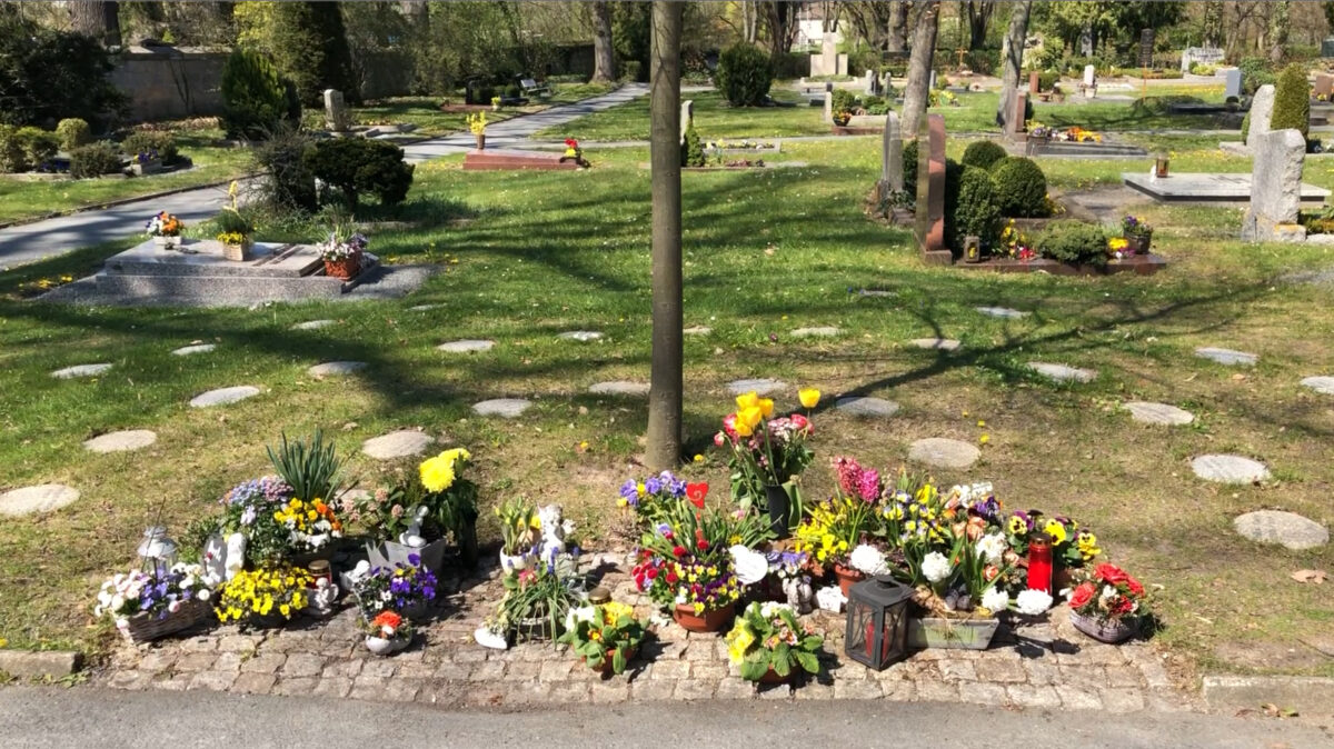Eine Friedhofsszene auf dem Bayreuther Stadtfriedhof zeigt Grabarten der Feuerbestattung mit bunten Blumenarrangements und Laternen vor einem Baum, umgeben von Grabsteinen und Grünpflanzen an einem sonnigen Tag.