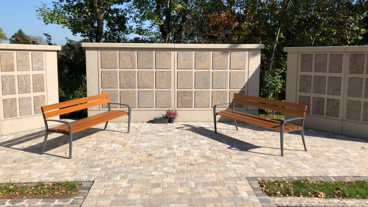 Zwei Holzbänke stehen sich auf einer gepflasterten Terrasse des Kolumbariums am Bayreuther Südfriedhof gegenüber, mit einer steinernen Kolumbariumsmauer im Hintergrund und Bäumen dahinter. Eine kleine Topfpflanze steht zwischen den Bänken an der Wand.