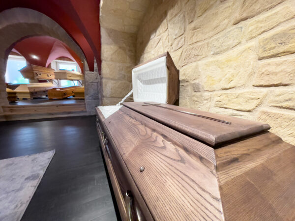 Nahaufnahme eines Holzsargs mit teilweise geöffnetem Deckel in einem Ausstellungsraum mit Steinwänden; im Hintergrund sind mehrere weitere Särge zu sehen.