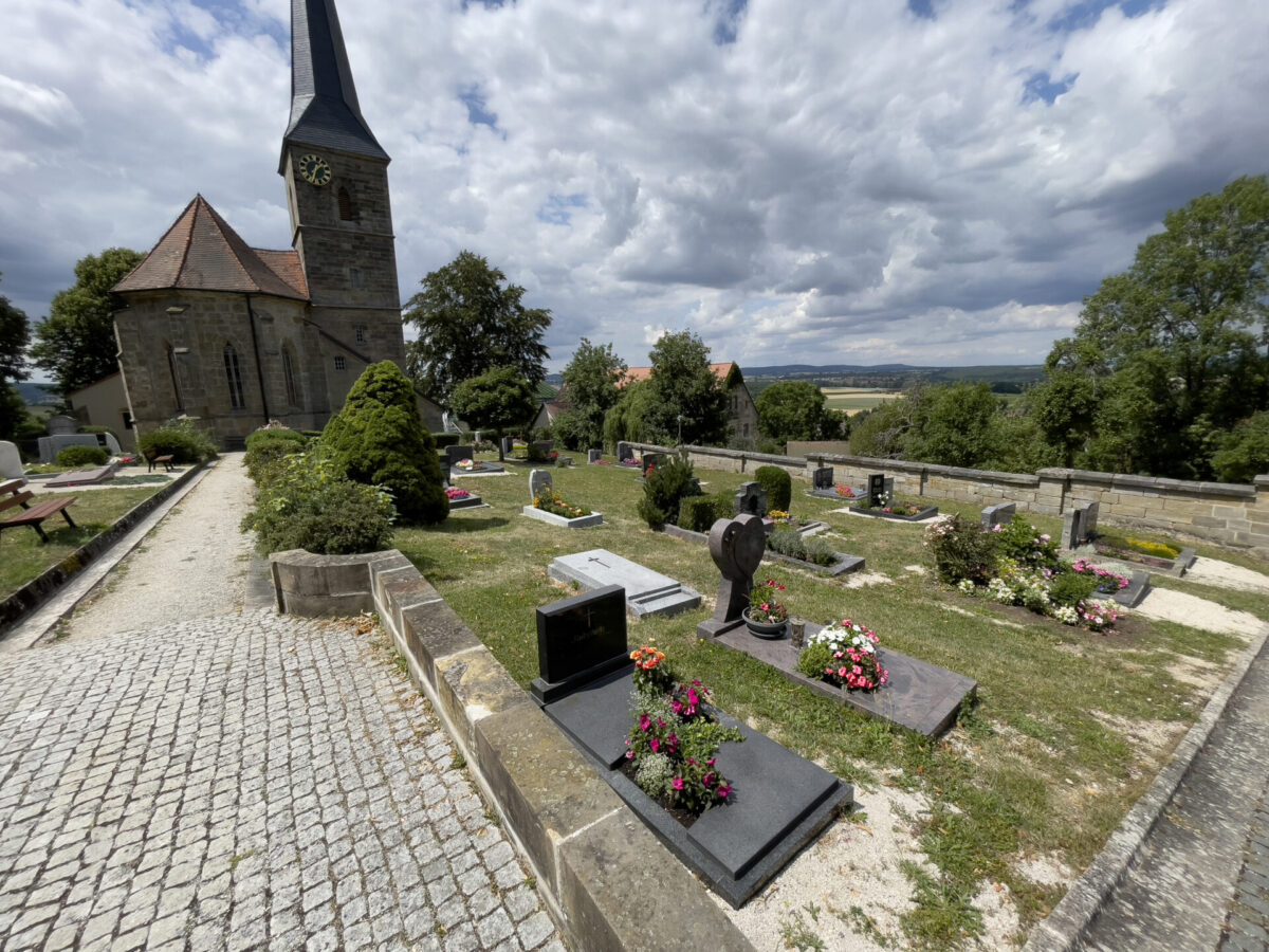 Der Friedhof Gesees mit Erdgräber liegt neben einer alten Steinkirche und einem Uhrenturm, mit Grabsteinen und bunten Blumenarrangements unter einem teilweise bewölkten Himmel, umgeben von Bäumen und fernen Feldern.