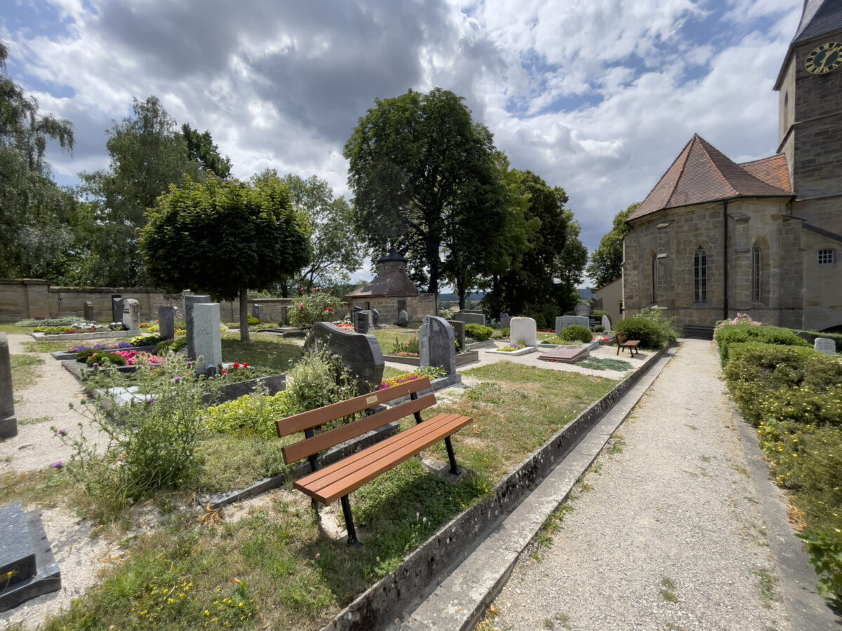 Ein Friedhof mit verschiedenen Grabsteinen (Ansicht Erdgräber), bunten Blumen und einer Holzbank im Vordergrund. Der Friedhof Gesees zeigt eine Kirche mit einem Glockenturm, großen Bäumen und einem bewölkten Himmel im Hintergrund.