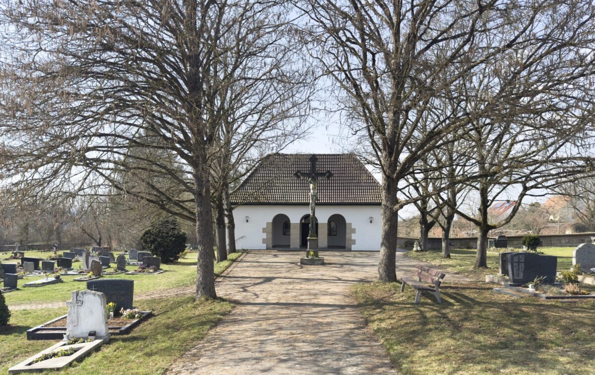 Eine kleine weiße Kapelle mit Ziegeldach steht am Ende eines von blattlosen Bäumen gesäumten Weges auf dem Erdgrab Friedhof Neudrossenfeld, umgeben von Grabsteinen und Bänken an einem klaren Tag.