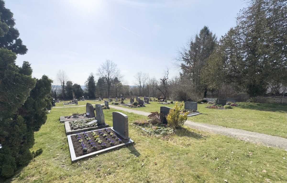 Ein friedlicher Friedhof mit Grabsteinen, Blumenbeeten und grünem Gras unter einem hellen, klaren Himmel. Bäume und Sträucher säumen den Friedhof Neudrossenfeld und werfen helle Schatten, während ein gepflasterter Weg durch das ruhige Gelände führt.