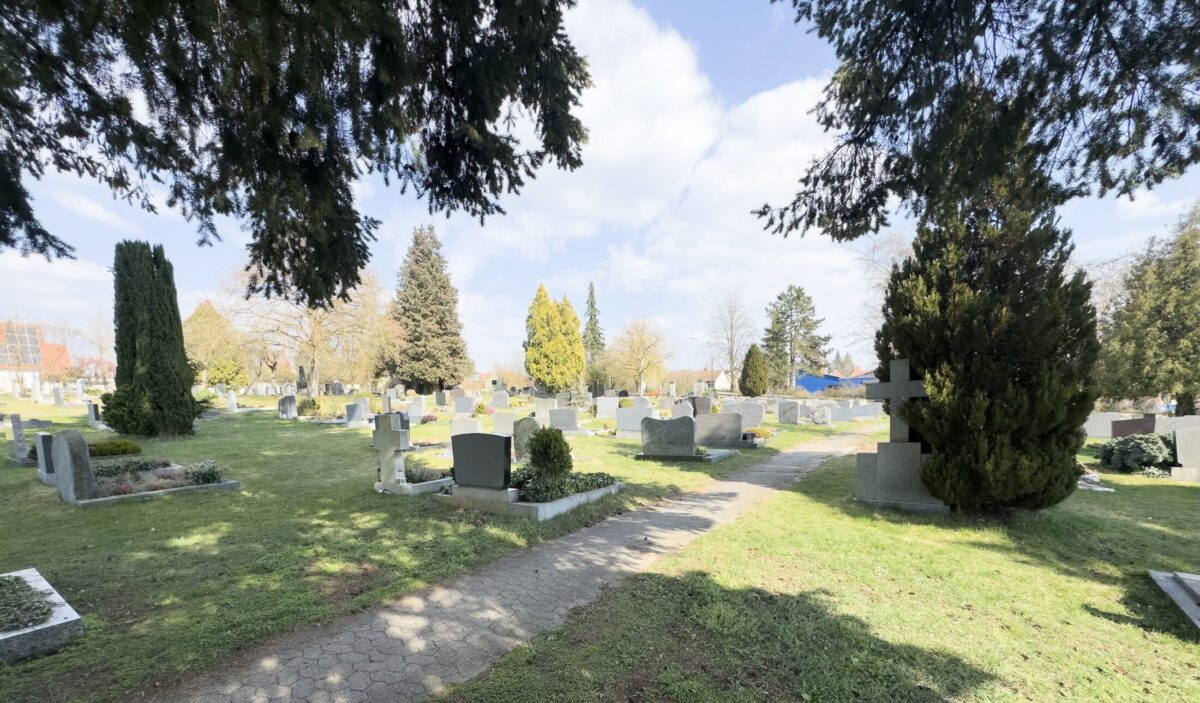 Ein friedlicher Friedhof, Friedhof Neudrossenfeld, mit verstreuten Grabsteinen auf einer Grasfläche, umgeben von Bäumen unter einem teilweise bewölkten Himmel. Ein gepflasterter Weg führt durch die Szene, mit Schatten von den Bäumen über dem Kopf.