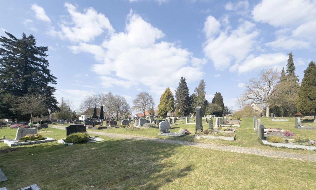 Ein friedlicher Friedhof, Friedhof Neudrossenfeld, mit Reihen von Grabsteinen und Denkmälern, grünem Gras und vereinzelten Bäumen unter einem teilweise bewölkten blauen Himmel. Einige Gräber haben Blumen oder kleine Sträucher neben sich.