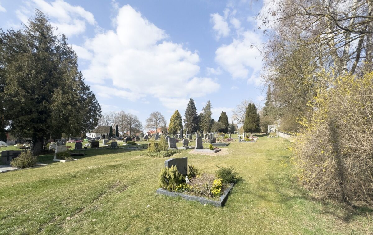 Eine friedliche Friedhofsszene auf dem Erdgrab Friedhof Neudrossenfeld mit verstreuten Grabsteinen auf einer grasbewachsenen Fläche, umgeben von hohen Bäumen und Büschen unter einem teilweise bewölkten Himmel.
