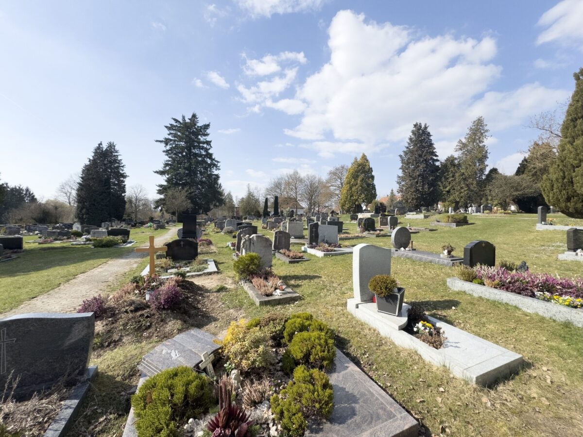 Ein Friedhof mit Reihen von Grabsteinen und Gräbern, einige mit Blumen und Pflanzen geschmückt, auf einer Wiese unter einem teilweise bewölkten Himmel mit Bäumen im Hintergrund auf dem Friedhof Neudrossenfeld.