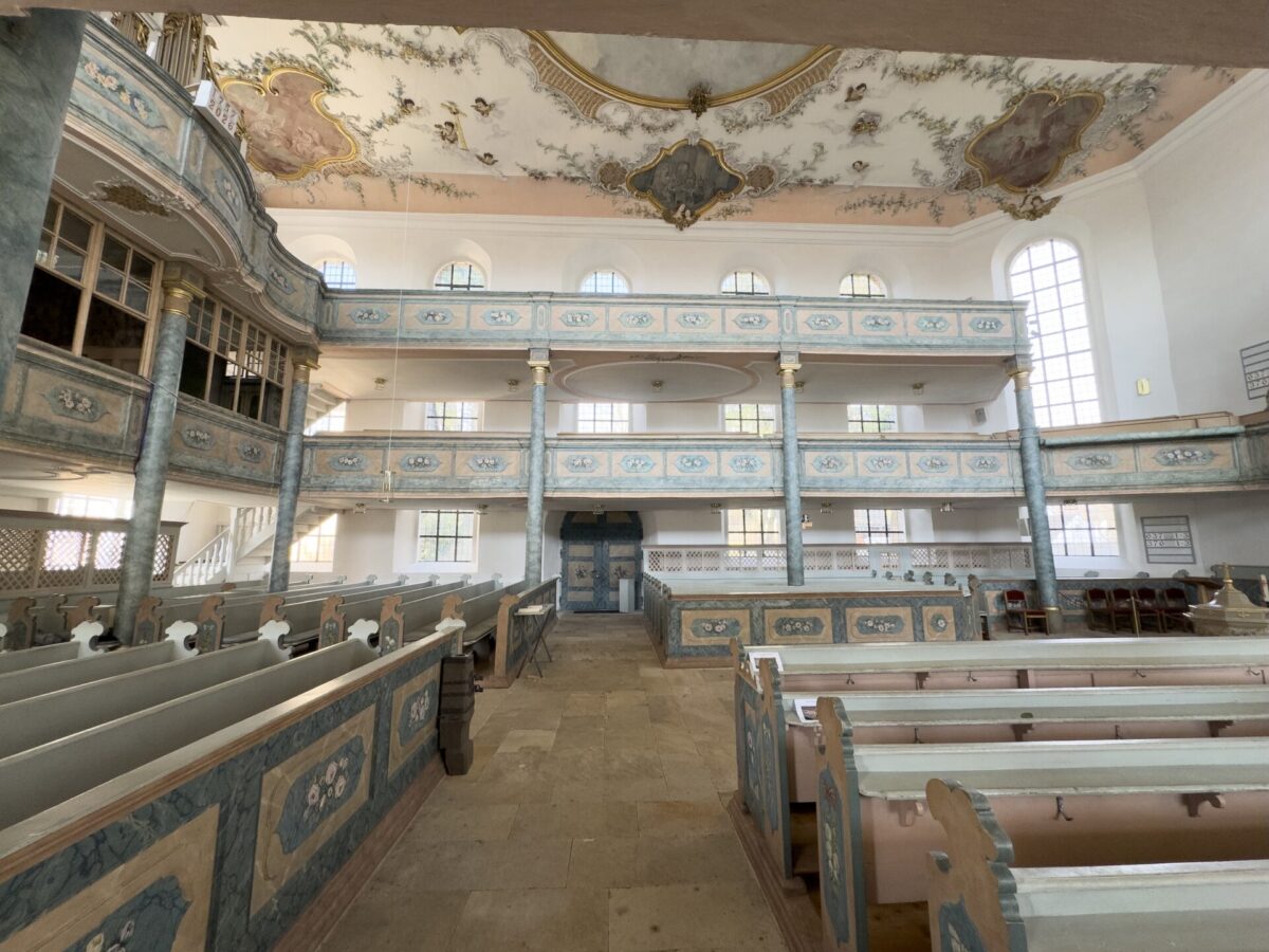 Innenraum einer kunstvollen, historischen Kirche in der Nähe des Friedhofs Neudrossenfeld mit pastellblauen und weißen Balkonen, verzierten Deckengemälden, großen Bogenfenstern und Reihen von Holzbänken. Natürliches Licht durchflutet den geräumigen, eleganten Saal.