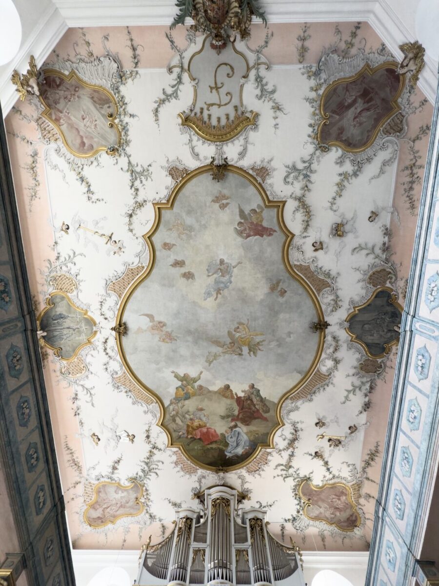 Verziertes Deckengemälde im Barockstil auf dem Friedhof Neudrossenfeld mit Engeln und Figuren in sanften Farben, umgeben von goldenen Zierleisten und floralen Mustern über einer großen Kapellenorgel.