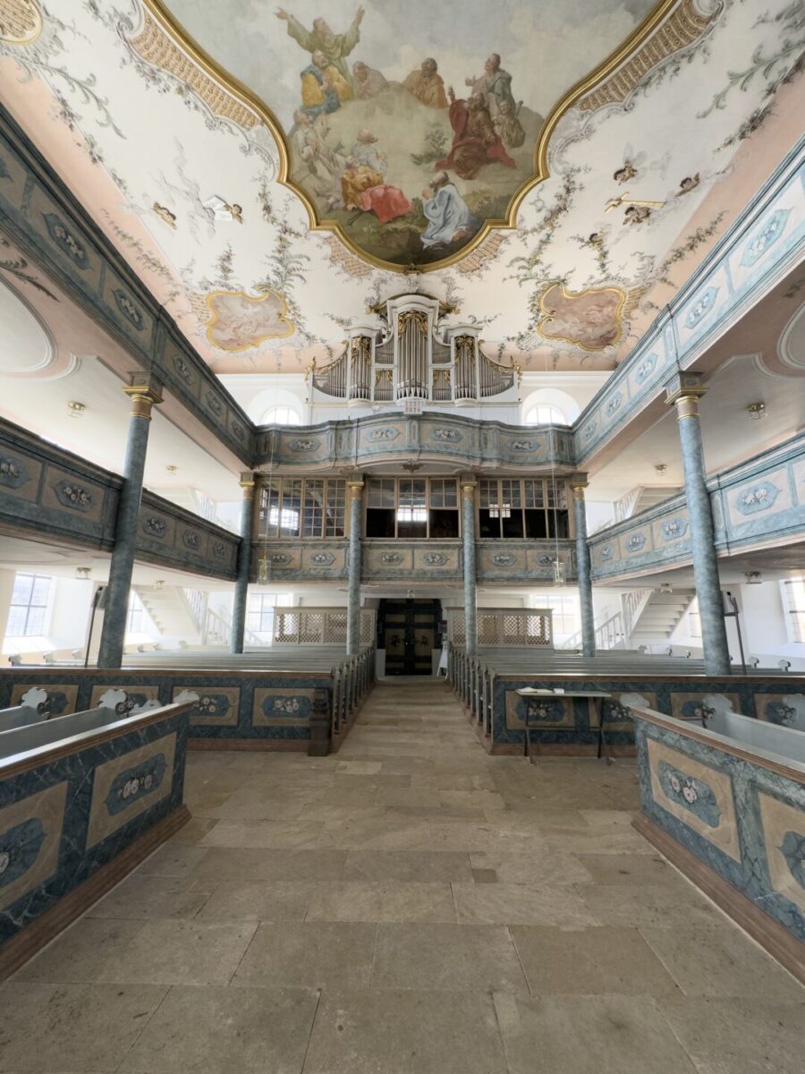 Das kunstvolle Kircheninnere des Friedhofs Neudrossenfeld mit dekorativen blauen Geländern, einer großen Pfeifenorgel über dem Eingang und einer kunstvoll bemalten Decke, die religiöse Szenen in Pastellfarben zeigt. Durch die hohen Fenster strömt natürliches Licht herein.