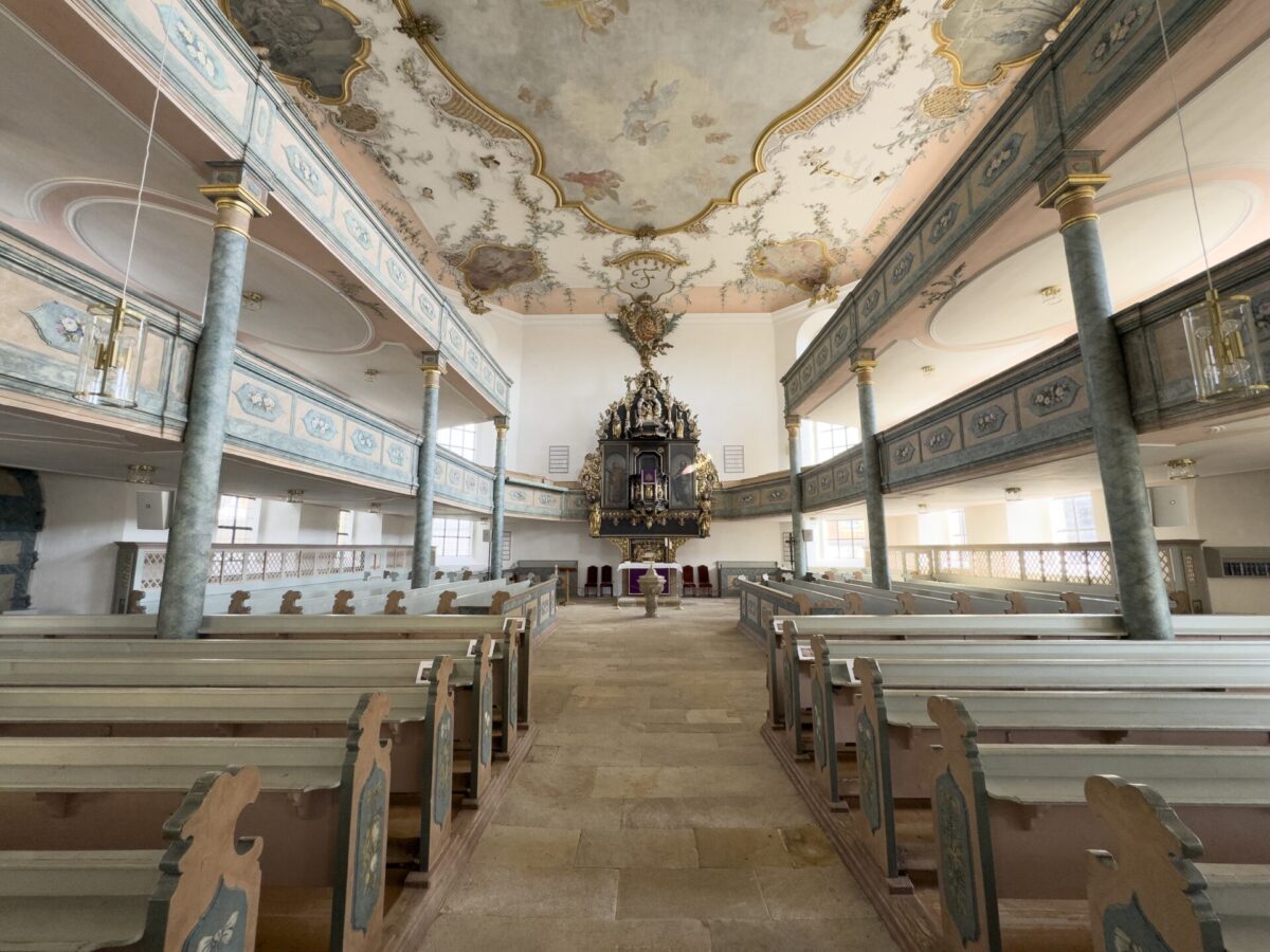 Ein weiter Blick in den kunstvollen Innenraum der Friedhofskirche Neudrossenfeld mit hellen Holzbänken, einer dekorativen Orgel an der Vorderseite, Marmorsäulen und einer bemalten Decke mit kunstvollen Mustern und sanftem Tageslicht, das den Raum erfüllt.