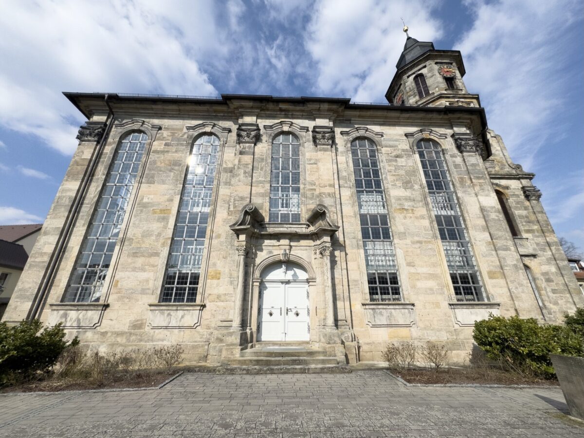 In der Nähe des friedlichen Friedhofs steht eine historische Steinkirche, die Kirche Neudrossenfeld, mit hohen Bogenfenstern, einer zentralen weißen Doppeltür und einem rechtsseitigen Turm vor einem blauen Wolkenhimmel.