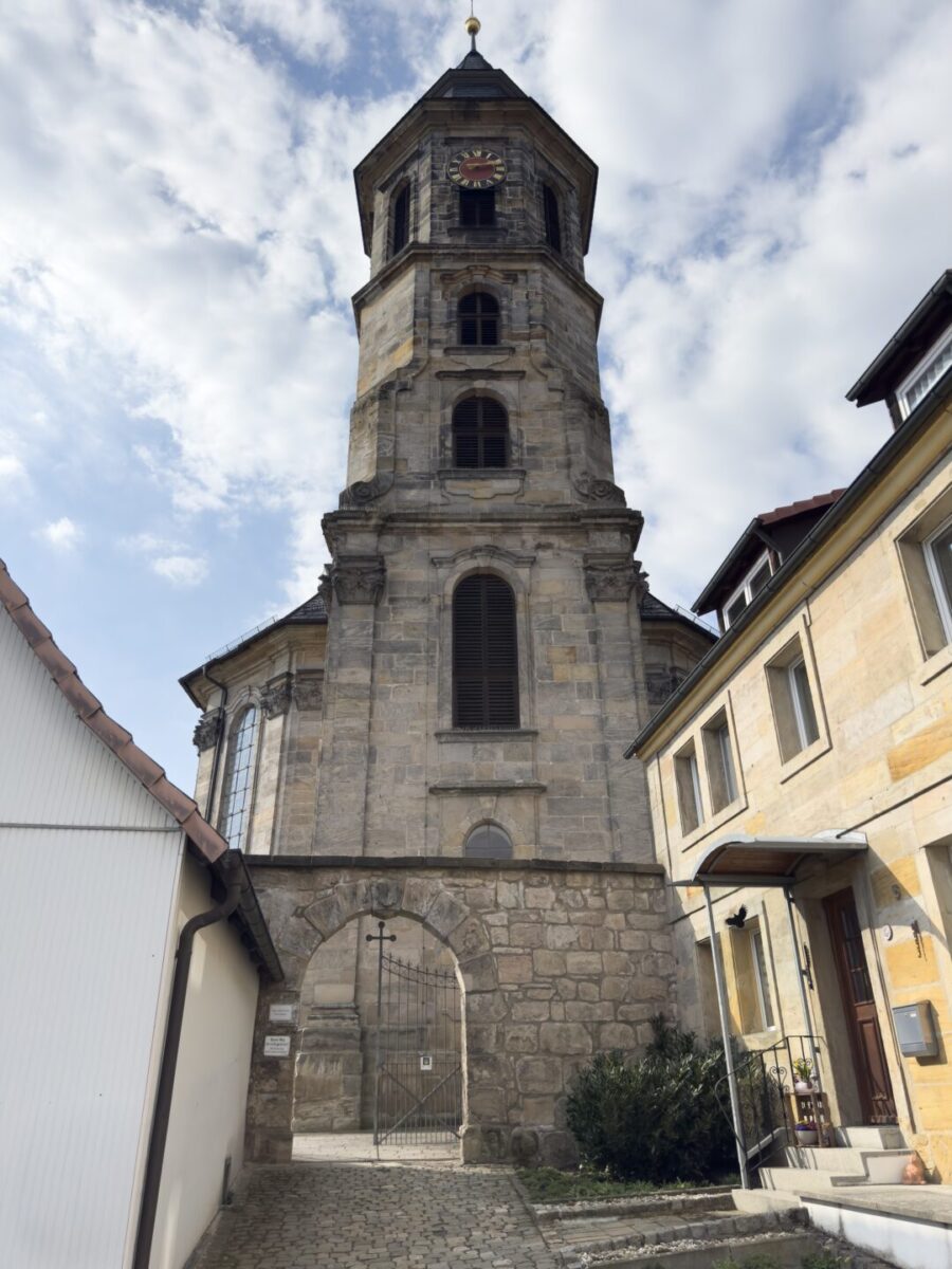 Ein hoher, historischer Steinkirchturm mit einer Uhr nahe der Spitze steht zwischen zwei Gebäuden unter einem teilweise bewölkten Himmel. Ein bogenförmiges Tor mit einem Eisenzaun führt zum Eingang unterhalb des Turms auf dem Friedhof Neudrossenfeld.