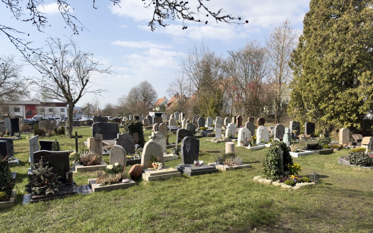 Auf dem Friedhof Neudrossenfeld befinden sich Reihen von Grabsteinen und Denkmälern, die teilweise mit Blumen und Pflanzen geschmückt sind, umgeben von Bäumen und Gras unter einem teilweise bewölkten Himmel. Im Hintergrund sind nahe gelegene Häuser zu sehen.