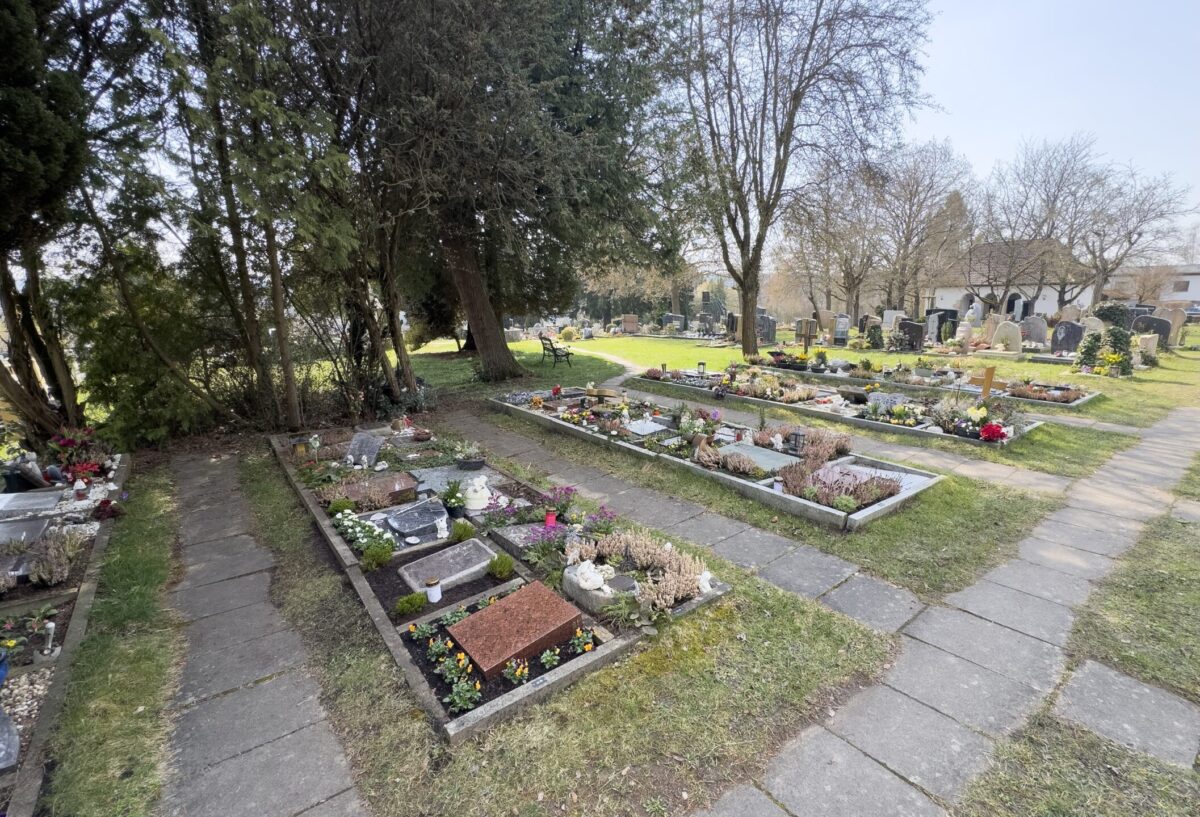 Auf dem Friedhof Neudrossenfeld befinden sich ordentlich angeordnete Gräber, die teilweise mit Blumen und Ornamenten geschmückt sind, umgeben von Bäumen und Gras, mit einem gepflasterten Weg und weiteren Grabsteinen im Hintergrund.
