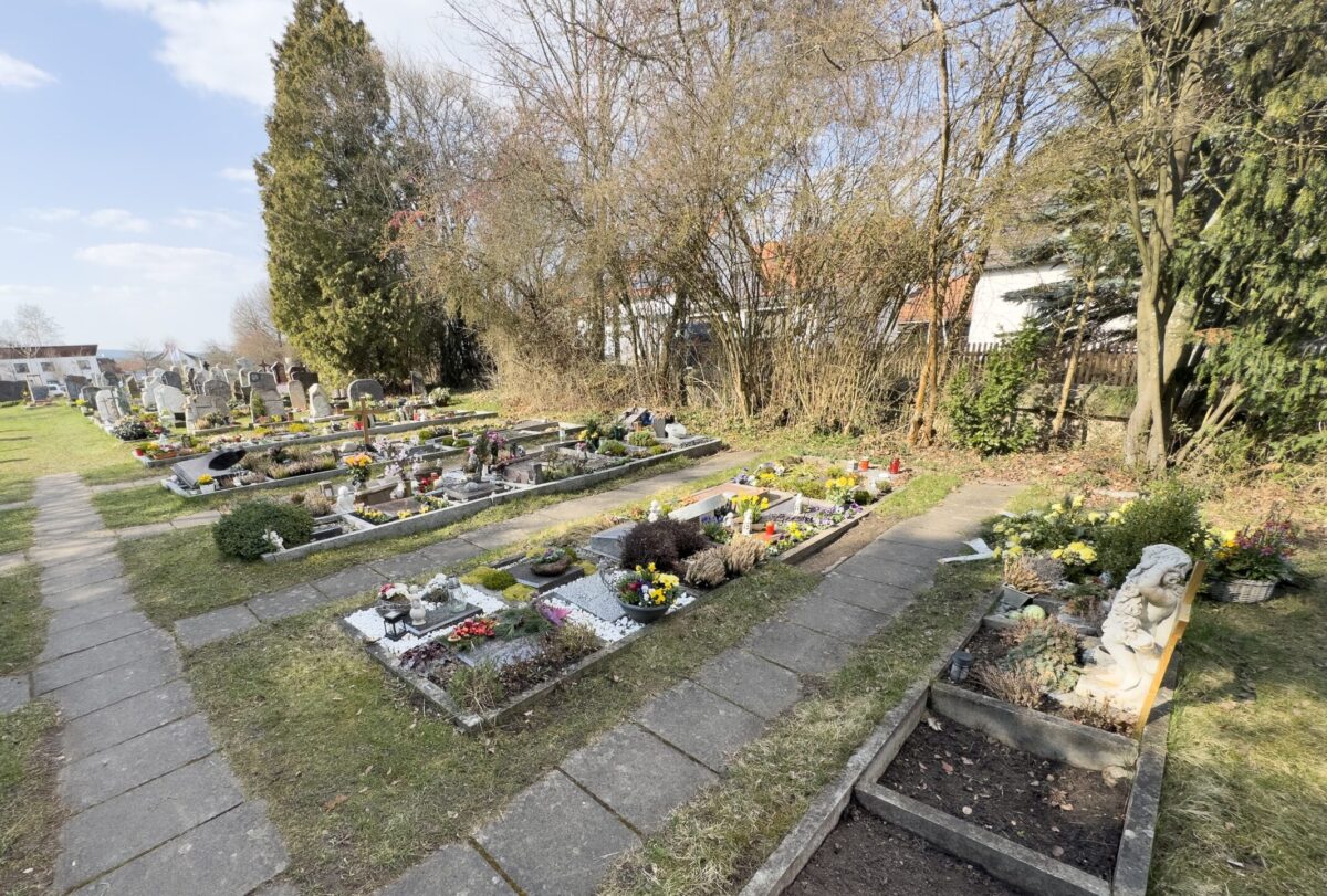 Ein sonnenbeschienener Friedhof mit ordentlich angelegten Gräbern, viele mit Blumen und kleinen Pflanzen geschmückt. Bäume und Sträucher säumen den Hintergrund des Friedhofs Neudrossenfeld, wo ein Steinweg entlang der Gräber verläuft.