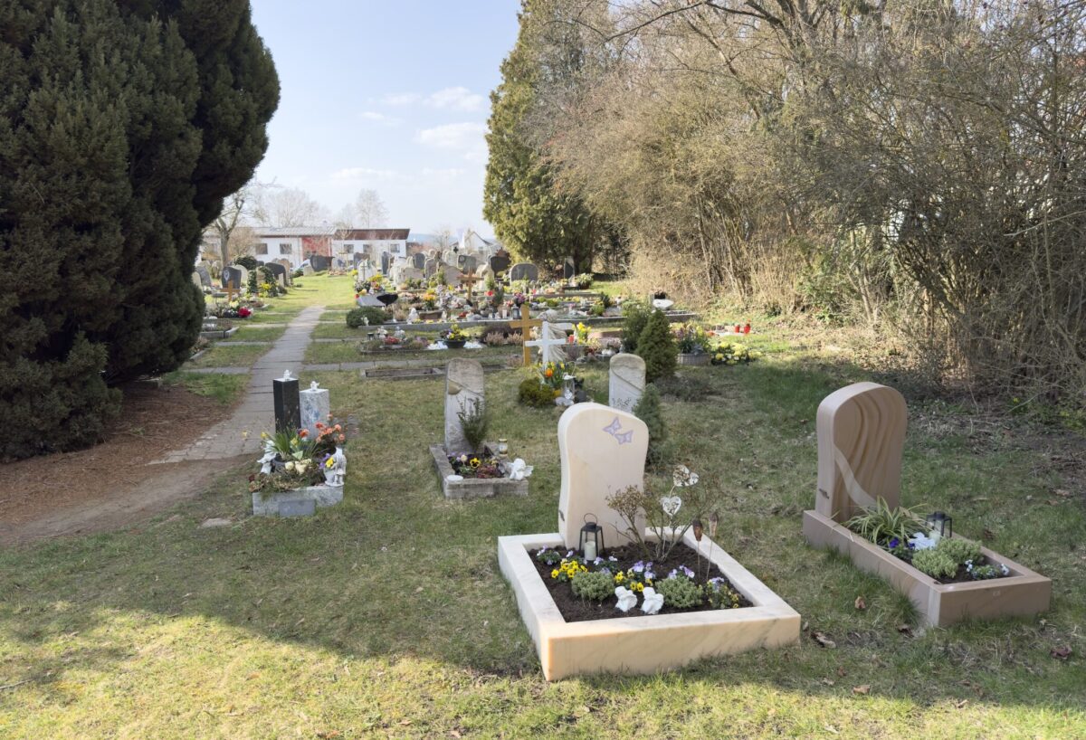Ein friedlicher Friedhof, Friedhof Neudrossenfeld, mit Grabsteinen und Blumenarrangements auf grasbewachsenen, von Bäumen und Sträuchern gesäumten Parzellen. Ein Fußweg führt zu den Häusern im Hintergrund unter einem teilweise bewölkten Himmel.