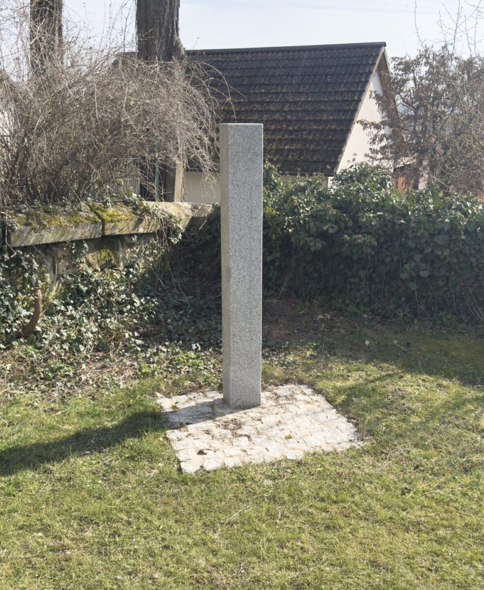Eine hohe, schmale, rechteckige Steinsäule steht auf einem kleinen quadratischen Fleck aus Pflastersteinen in einem grasbewachsenen Hof auf dem Friedhof Neudrossenfeld, umgeben von Büschen, Efeu und einem Holzzaun mit Häusern im Hintergrund.