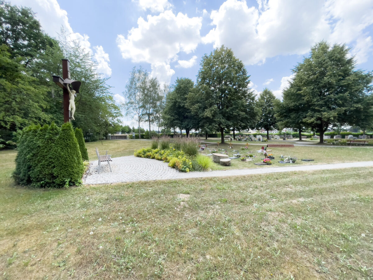 Ein friedlicher Friedhof mit Urnengräbern mit liegendem Stein am Waldfriedhof Eckersdorf, einem großen hölzernen Kruzifix, Bänken, Gräbern mit Blumen und alten Bäumen unter einem teilweise bewölkten Himmel.