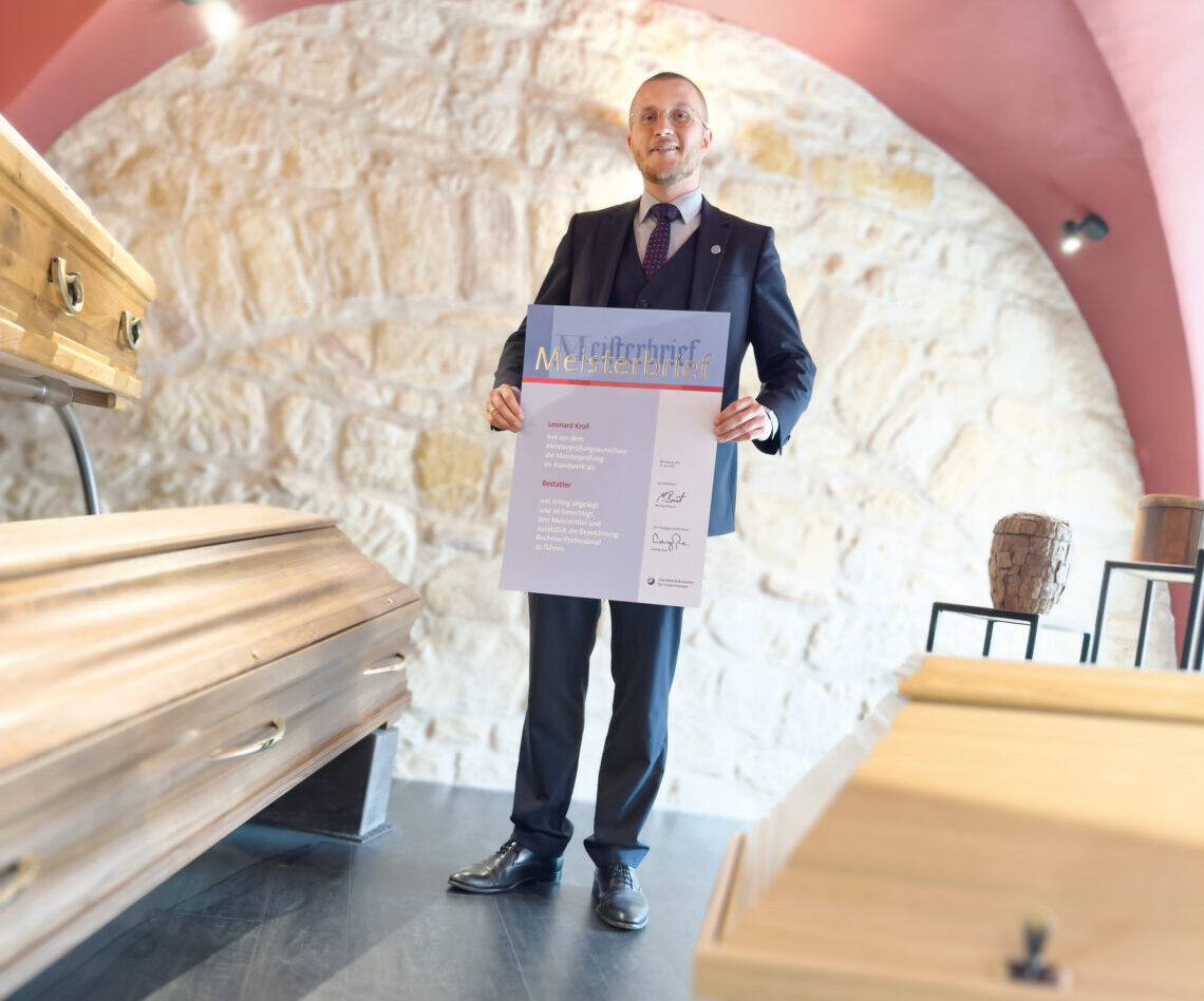Ein Leonard Kroll im Anzug hält das Zertifikat des Bestattermeisters in der Hand, während er in einem Raum mit Steinwänden, einer rosa Decke und mehreren ausgestellten Holzsärgen steht.