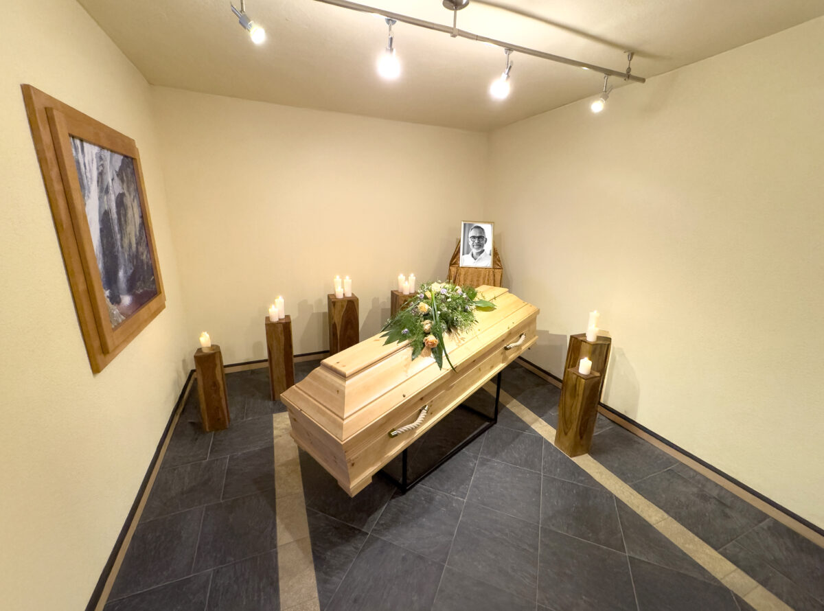 Ein schlichter Raum der Zeit mit einem blumengeschmückten Holzsarg, brennenden Kerzen auf Ständern rundherum und einem gerahmten Schwarz-Weiß-Porträt einer Person auf dem Sarg. Ein Bild hängt an der beigen Wand links von Himml Bestattungen.