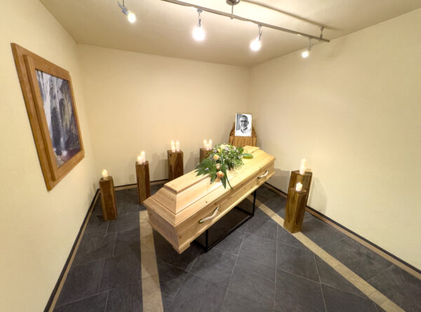 Ein schlichter Raum der Zeit mit einem blumengeschmückten Holzsarg, brennenden Kerzen auf Ständern rundherum und einem gerahmten Schwarz-Weiß-Porträt einer Person auf dem Sarg. Ein Bild hängt an der beigen Wand links von Himml Bestattungen.