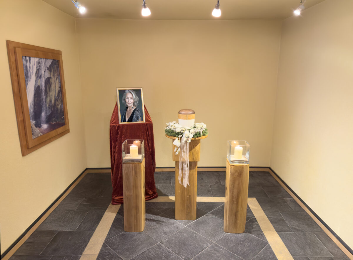 Ein Gedenkraum mit dem gerahmten Foto einer lächelnden Frau auf einem drapierten Ständer, einem Kranz mit einer Kerzenurne auf einem zentralen Sockel und zwei umliegenden Sockeln mit je einer brennenden Kerze. Sanfte Beleuchtung erhellt den Raum der Zeit bei Himml Bestattungen.