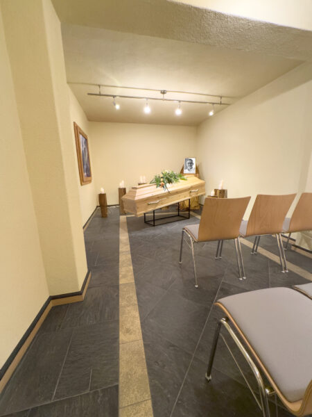 In einer kleinen Grabkapelle von Himml Bestattungen, genannt Raum der Zeit, steht ein Holzsarg, der mit Blumen und Kerzen geschmückt ist. Leere Holzstühle stehen daneben, und ein gerahmtes Schwarz-Weiß-Foto ist unter sanfter Deckenbeleuchtung zu sehen.