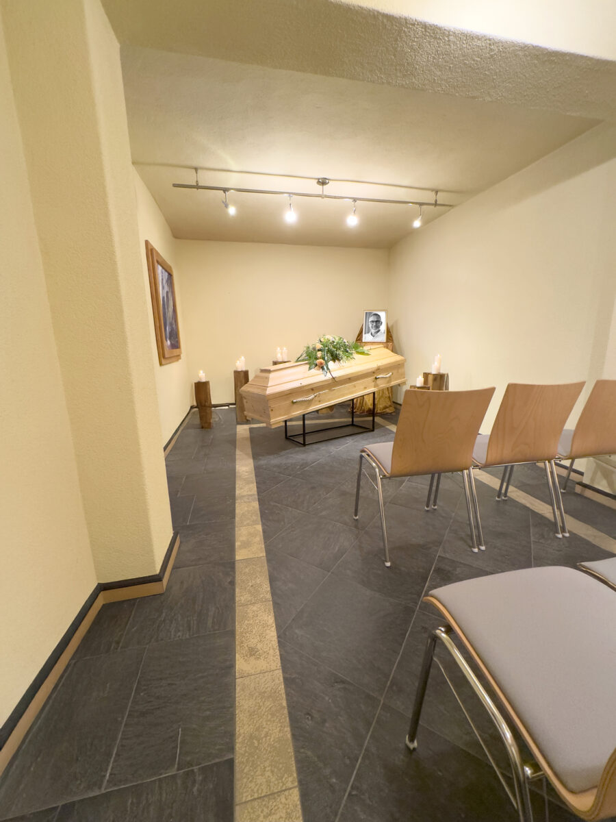 In einer kleinen Grabkapelle von Himml Bestattungen, genannt Raum der Zeit, steht ein Holzsarg, der mit Blumen und Kerzen geschmückt ist. Leere Holzstühle stehen daneben, und ein gerahmtes Schwarz-Weiß-Foto ist unter sanfter Deckenbeleuchtung zu sehen.