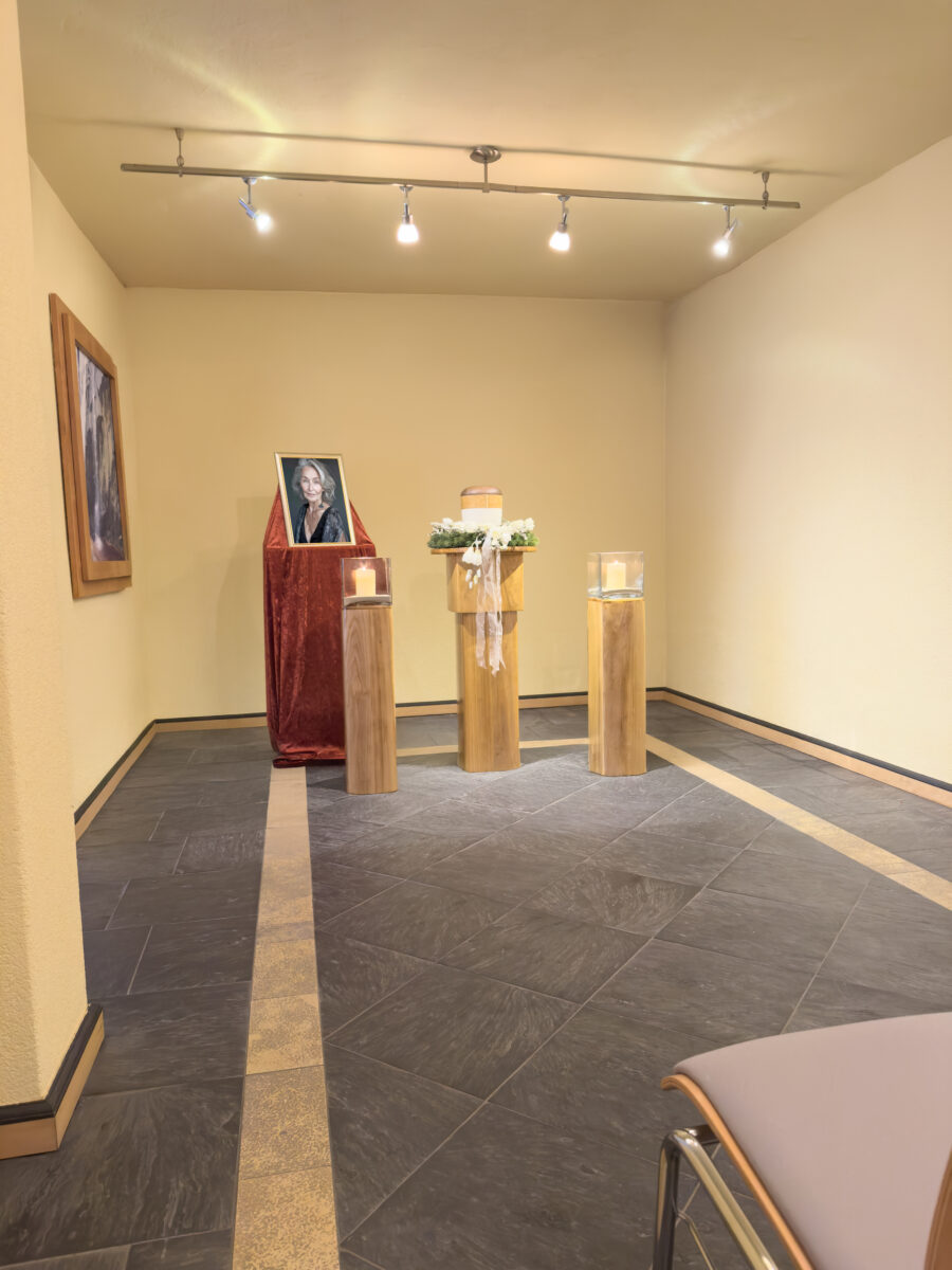 Eine Trauerfeier von Raum der Zeit Himml Bestattungen mit einem gerahmten Porträt auf rotem Samt, einer weißen, mit Blumen geschmückten Urne auf einem Holzsockel, zwei großen Kerzen und sanfter Deckenbeleuchtung in einem ruhigen, beigen Raum.