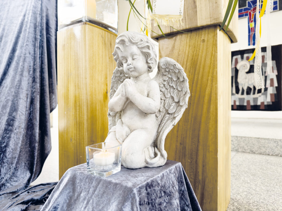 Eine kleine Engelsstatue mit gefalteten Händen kniet auf einem stoffbezogenen Sockel neben einer brennenden Kerze in einem Glashalter. So entsteht eine friedliche Szene, wie man sie oft bei einer Trauerfeier sieht, inmitten von Holz- und Stoffdekorationen in einem hellen Innenraum.