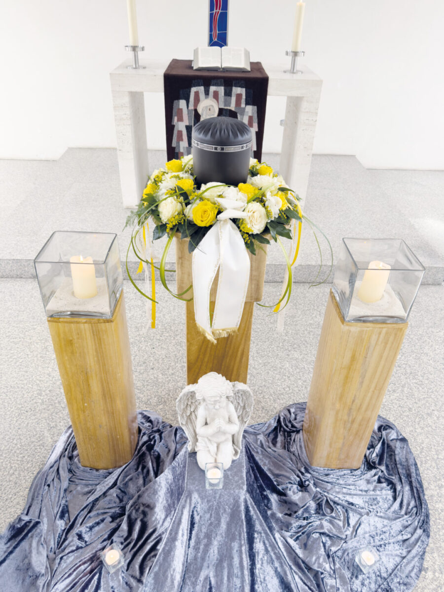 Eine schwarze Urne ruht auf einem mit gelben und weißen Blumen und einem Band geschmückten Sockel, flankiert von Kerzen auf Holzständern, darunter eine weiße Engelsfigur und ein drapiertes silbernes Tuch - eine elegante Kulisse für eine Trauerfeier mit einem Altar im Hintergrund.