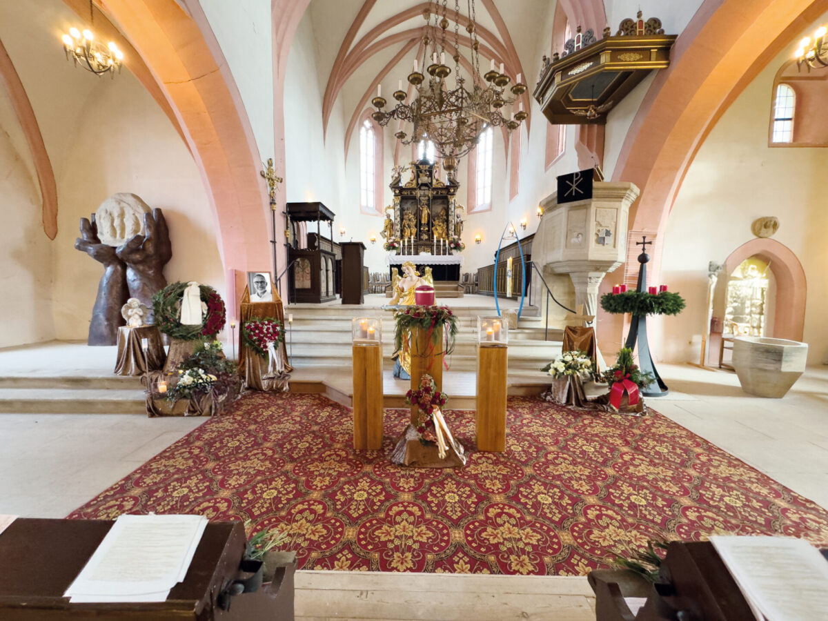 Ein für eine Trauerfeier geschmückter Kirchenaltar mit Kerzen, Kränzen und Blumenschmuck. Der Raum weist gewölbte Decken, einen rot gemusterten Teppich, verzierte Kronleuchter und Kirchenbänke mit Notenständern im Vordergrund auf.