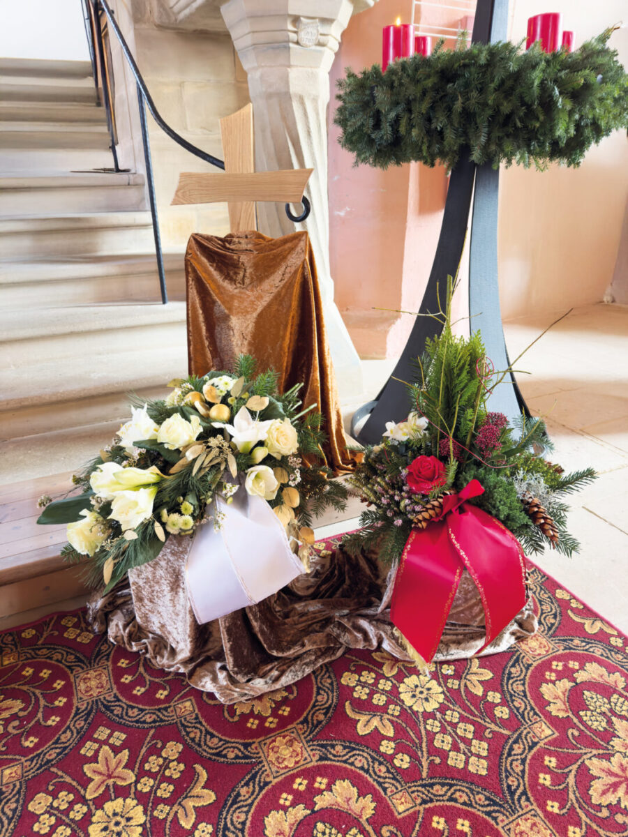 Ein Gesteck mit weißen Blumen und einer weißen Schleife und ein weiteres mit roten Rosen und einer roten Schleife, ein Holzkreuz, drapierte Stoffe und ein Adventskranz mit vier roten Kerzen in der Kirche für eine Trauerfeier.
