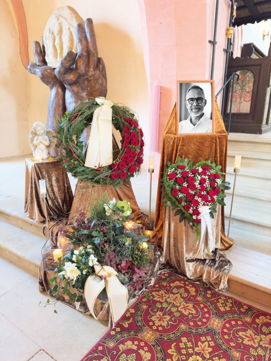 Ein Gedenkbild für eine Trauerfeier zeigt ein gerahmtes Schwarz-Weiß-Foto eines Mannes, Blumenkränze, Kerzen und eine Skulptur mit großen, betenden Händen, die alle sorgfältig auf einem Bronzetuch in einer Kirche arrangiert sind.