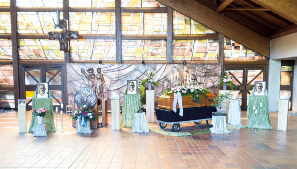 Ein mit Blumen geschmückter Holzsarg steht in einer sonnendurchfluteten Kapelle mit Buntglas. In der Nähe stehen vier Gedenkporträts, die jeweils von Blumenarrangements umgeben sind. Die Szene erinnert an eine Beerdigung oder einen Gedenkgottesdienst.