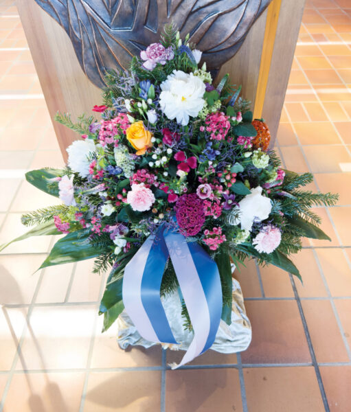 Ein großes, farbenfrohes Blumenarrangement mit weißen, rosafarbenen, lila und gelben Blumen, akzentuiert mit Grünzeug und einem blau-weißen Band, auf einem gefliesten Boden vor einem hölzernen Hintergrund - ideal für eine Trauerfeier mit Sarg.