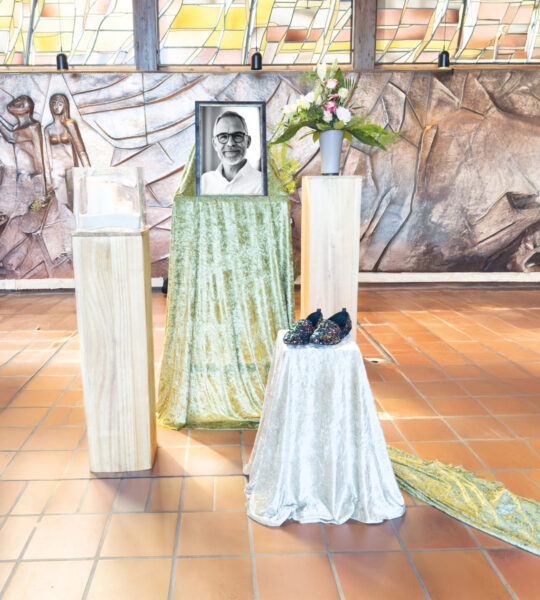 Eine Gedenkstätte für eine Trauerfeier zeigt das Schwarz-Weiß-Porträt eines lächelnden Mannes auf einem drapierten Sockel. Kerzen, Schuhe und Blumen sind neben einem symbolischen Sarg in einem gekachelten Raum mit kunstvollen Wandschnitzereien angeordnet.