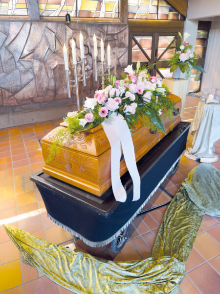 Ein hölzerner Sarg, der mit einem großen Blumenarrangement und weißem Band geschmückt ist, wird während einer Trauerfeier in der Kapelle aufgestellt. Hinter dem Sarg stehen brennende Kerzen, und auf dem Boden davor ist grüner Stoff drapiert.