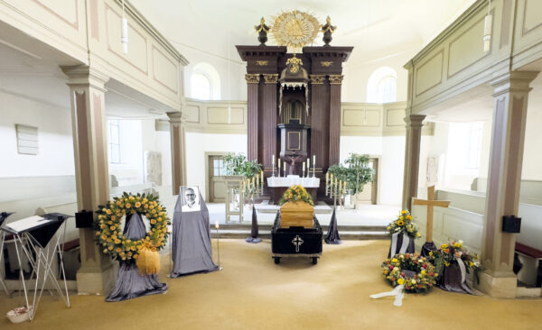 Eine Trauerfeier in einer Kirche, mit einem Sarg in der Mitte, umgeben von Blumenkränzen, einem gerahmten Foto des Verstorbenen und Kerzen auf dem Altar im Hintergrund.