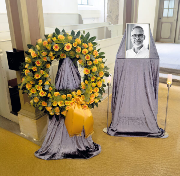 Eine Trauerfeier mit einem großen gelben Blumenkranz und einem gerahmten Schwarz-Weiß-Foto eines älteren Mannes mit Brille, beide auf mit silbernem Stoff bezogenen Ständern, daneben zwei brennende Kerzen.