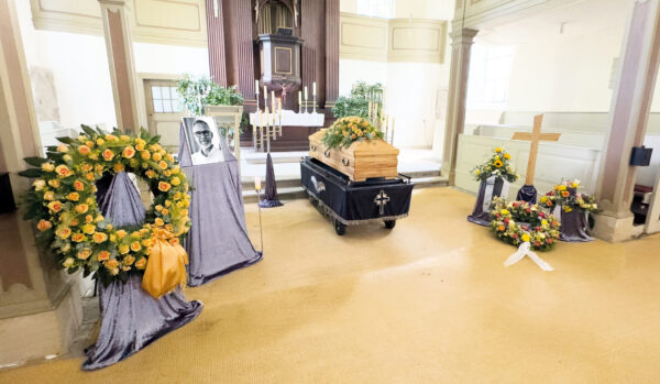 Ein hölzerner Sarg, umgeben von Blumenkränzen und einem Kreuz in einer Kirche. Während der Trauerfeier wird neben dem Sarg ein Schwarz-Weiß-Porträt gezeigt, während im Hintergrund in der Nähe des Altars Kerzen brennen.