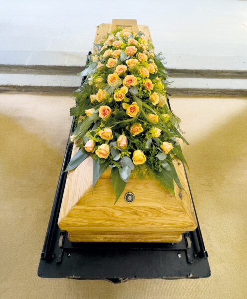 Ein hölzerner Sarg, geschmückt mit einem großen Strauß gelber und orangefarbener Rosen, steht in einem hellen Raum, der wahrscheinlich für eine Trauerfeier vorbereitet ist.