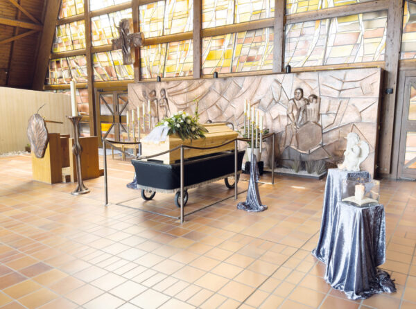Ein Sarg mit Blumen ruht auf einem Ständer in einem großzügigen, lichtdurchfluteten Raum mit Buntglasfenstern und Bronzereliefs. Kerzen und Dekoständer umgeben den Sarg - eine würdevolle Atmosphäre für die Trauerfeier.