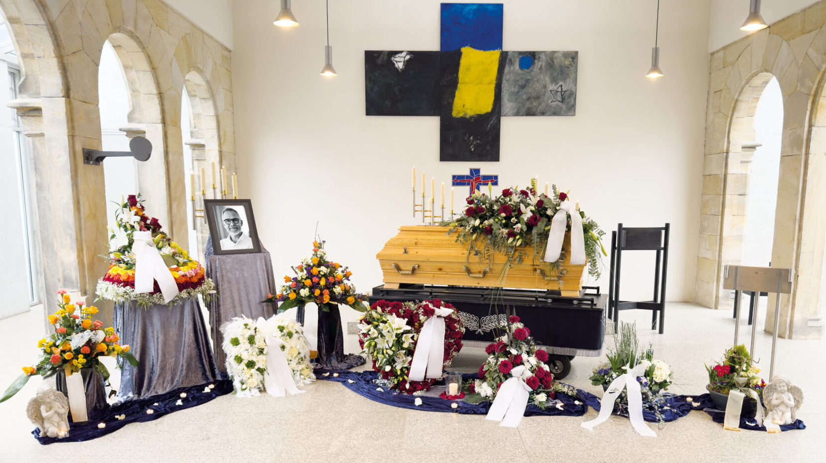Eine Beerdigungssituation mit einem blumengeschmückten Holzsarg, umgeben von Kränzen und Blumenarrangements. Links steht ein gerahmtes Schwarz-Weiß-Foto, darunter ein großes Kreuz und ein abstraktes Kunstwerk an der Wand.
