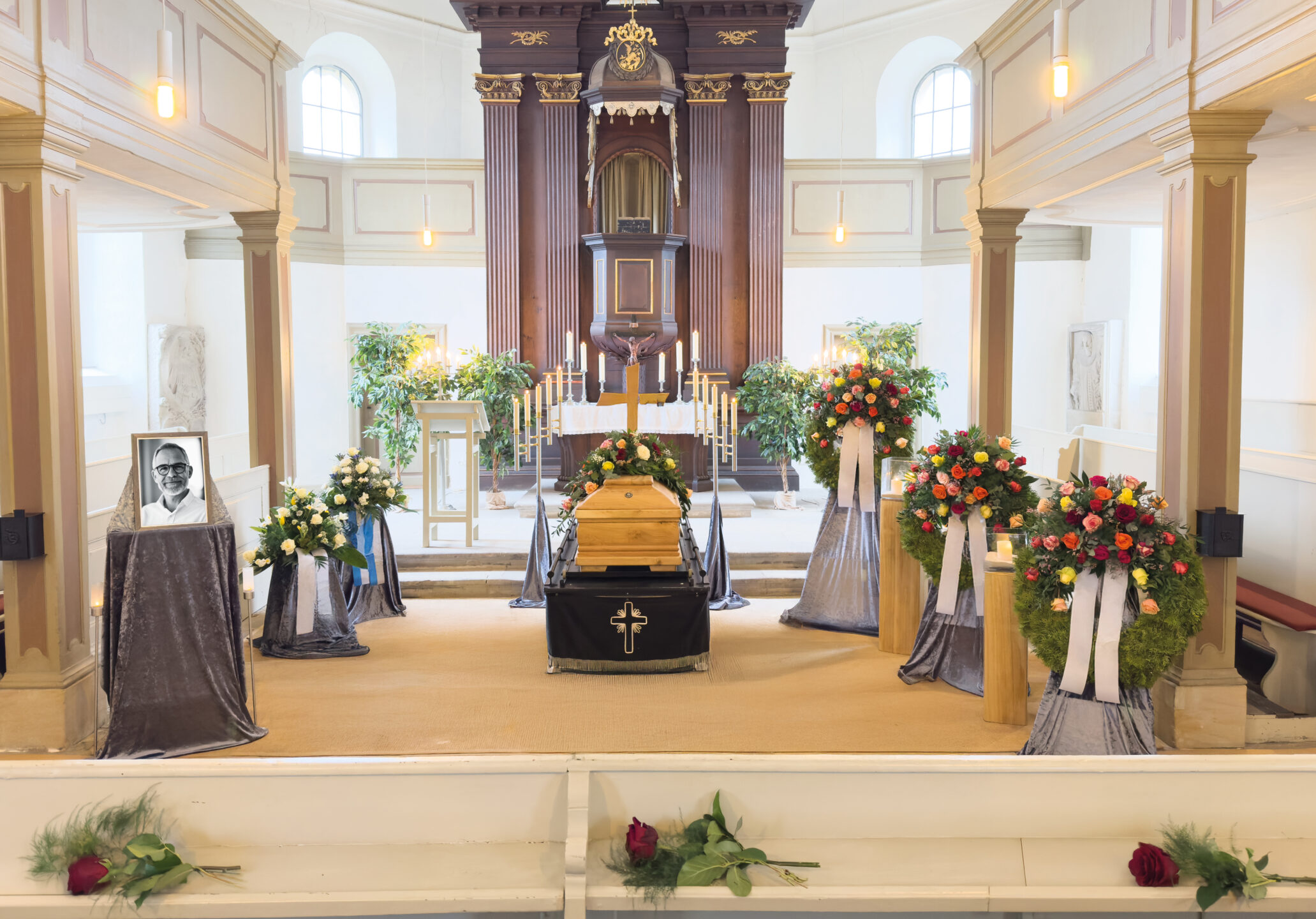 Ein Kirchenraum für eine Beerdigung mit einem Sarg mit Kreuz, Blumenkränzen, einem gerahmten Foto auf einem Ständer, Kerzen und Rosen auf den weißen Kirchenbänken. Die Umgebung ist hell mit natürlichem Licht.