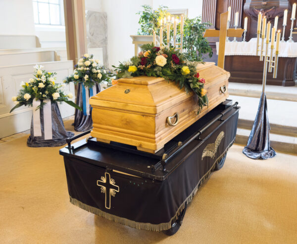 Ein hölzerner Sarg mit einem Blumengesteck ruht auf einem Wagen, der mit einem schwarzen Tuch mit Kreuzmotiv bedeckt ist, in einer mit Blumen und brennenden Kerzen für die Trauerfeier geschmückten Kirche.