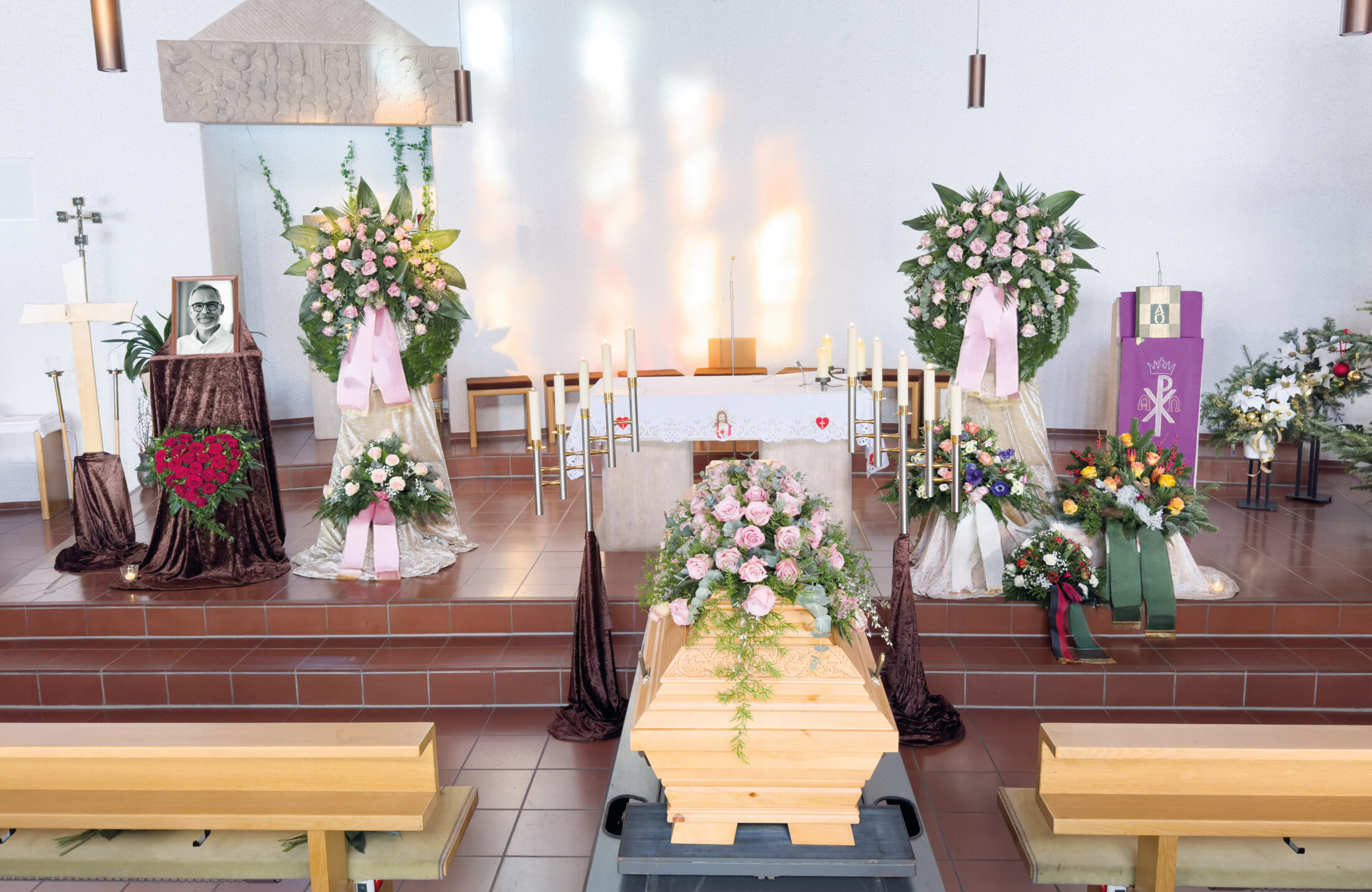 Ein mit rosa Rosen geschmückter Holzsarg steht in einer Kirche, umgeben von großen Blumenarrangements, Kerzen und einem gerahmten Schwarz-Weiß-Foto, das auf eine Beerdigung oder eine Gedenkfeier hinweist.