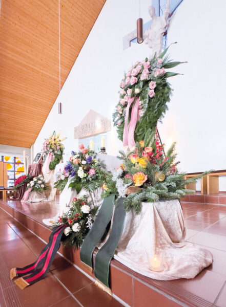 Ein Beerdigungsaltar in einer Kirche zeigt Blumengestecke, Kränze mit Bändern und Kerzen auf drapierten weißen Tüchern. Während der Trauerfeier mit Sarg ist eine Jesusstatue über dem Altar vor einer weißen Wand zu sehen.