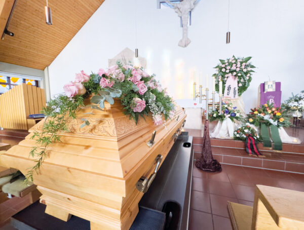 Ein hölzerner Sarg, der mit rosa und weißen Blumen geschmückt ist, steht in einer Kirche, umgeben von Blumenarrangements und Kerzen. Die Szenerie deutet auf eine Trauerfeier oder einen Gedenkgottesdienst hin.